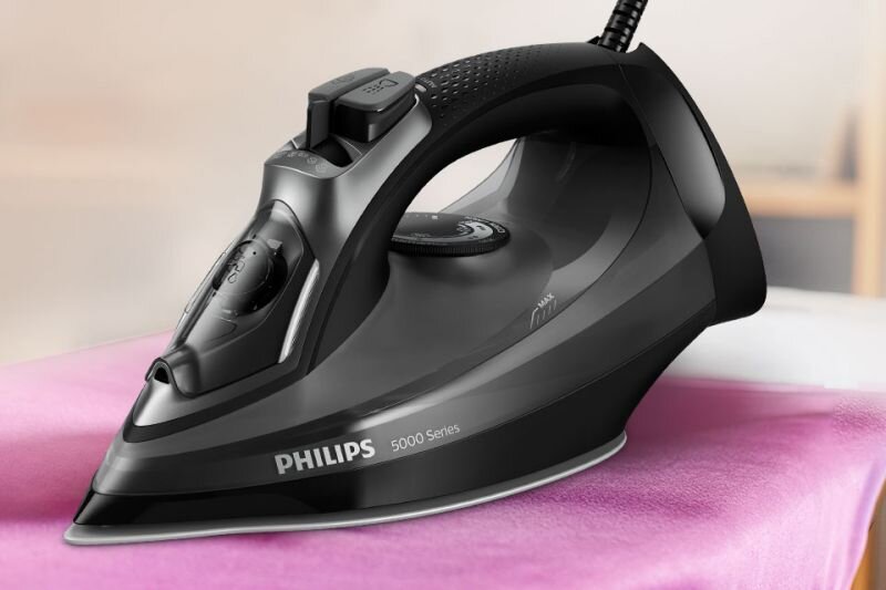Czarne żelazko z napisem \'PHILIPS 5000 Series\' stoi na różowym materiale. W tle widoczne są rozmyte elementy wnętrza.