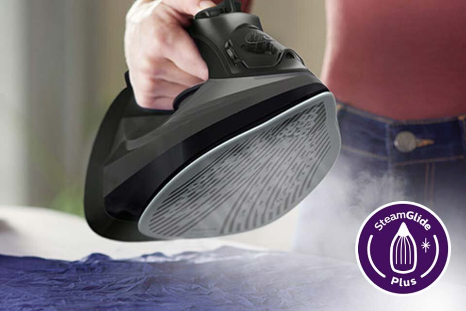 Osoba trzyma żelazko z widoczną stopą grzewczą, z której wydobywa się para. Obok znajduje się fioletowe logo z napisem \'SteamGlide Plus\'.