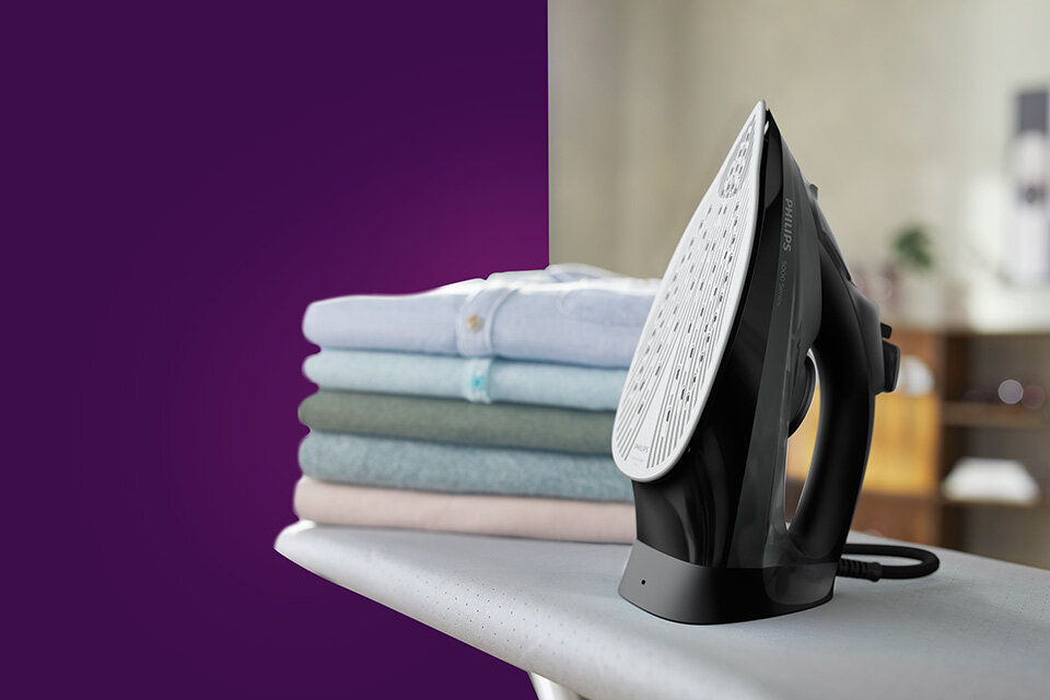 Na desce do prasowania stoi czarne żelazko z widoczną stopą, obok znajduje się stos złożonych ubrań. W tle widoczny jest rozmyty fragment wnętrza pomieszczenia.