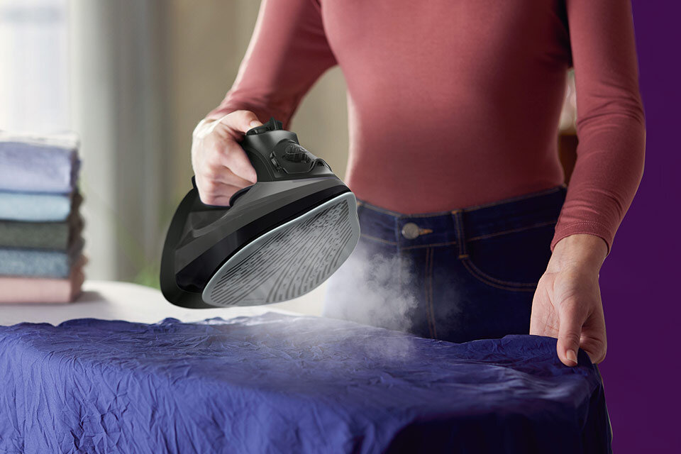 Osoba prasuje niebieski materiał za pomocą żelazka, z którego wydobywa się para. W tle widoczne są złożone ręczniki w różnych kolorach.