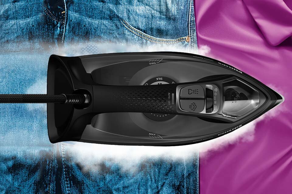 Czarne żelazko z widocznym uchwytem i przyciskami, emitujące parę, znajduje się na tle niebieskiego dżinsu i fioletowej tkaniny. Na żelazku widoczny jest tekst: \'MAX\', \'SILK\', \'MIN\'.