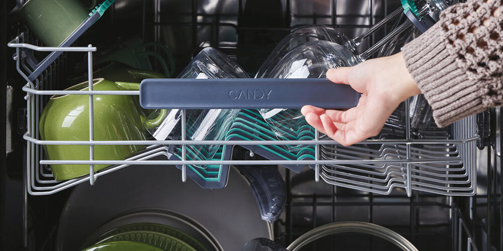Osoba trzyma uchwyt kosza zmywarki z napisem \'CANDY\'. Wewnątrz znajdują się naczynia, w tym zielone kubki i przezroczyste szklanki.