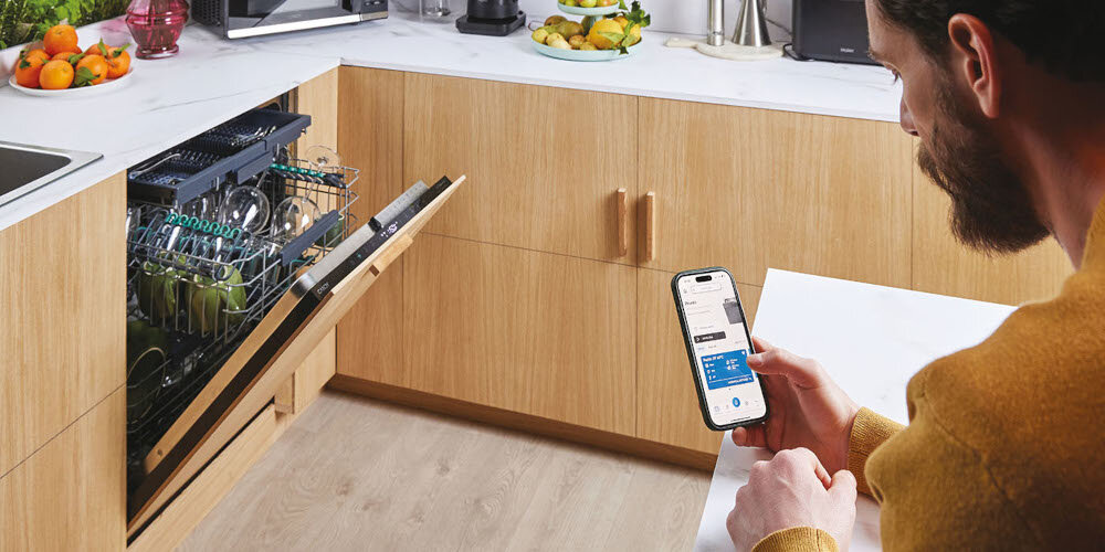 Osoba trzymająca smartfon z aplikacją, siedzi przy blacie kuchennym obok otwartej zmywarki z widocznymi naczyniami. W tle widać drewniane szafki kuchenne i różne przedmioty na blacie.