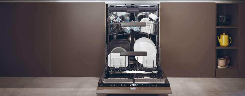 Otwarta zmywarka z widocznymi naczyniami w środku, umieszczona w zabudowie kuchennej. Na panelu zmywarki widoczne jest logo \'Haier\'.