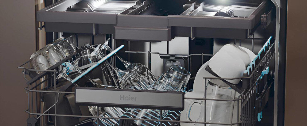 Wnętrze zmywarki z widocznymi naczyniami, takimi jak szklanki i talerze, ułożonymi w koszach. Na froncie zmywarki widoczne jest logo \'Haier\'.