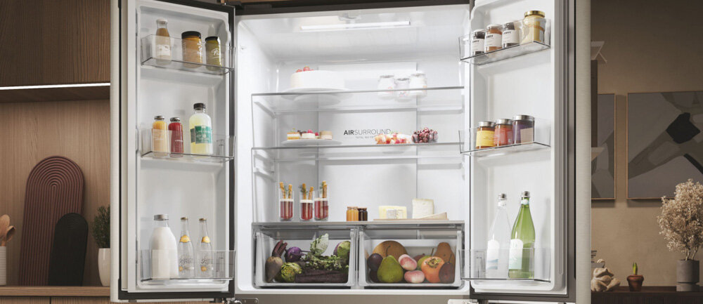 Otwarta lodówka z widocznymi półkami wypełnionymi różnorodnymi produktami spożywczymi, takimi jak butelki, słoiki, owoce i warzywa. Na tylnej ściance widoczny jest napis \'AIR SURROUND\'.