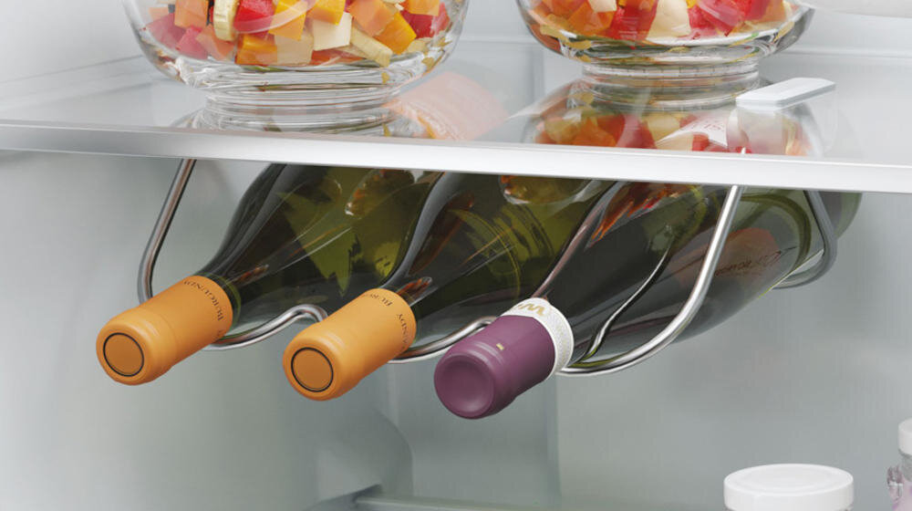 Wnętrze lodówki z trzema butelkami wina umieszczonymi na metalowym stojaku oraz dwoma szklanymi miskami z pokrojonymi owocami na półce powyżej.