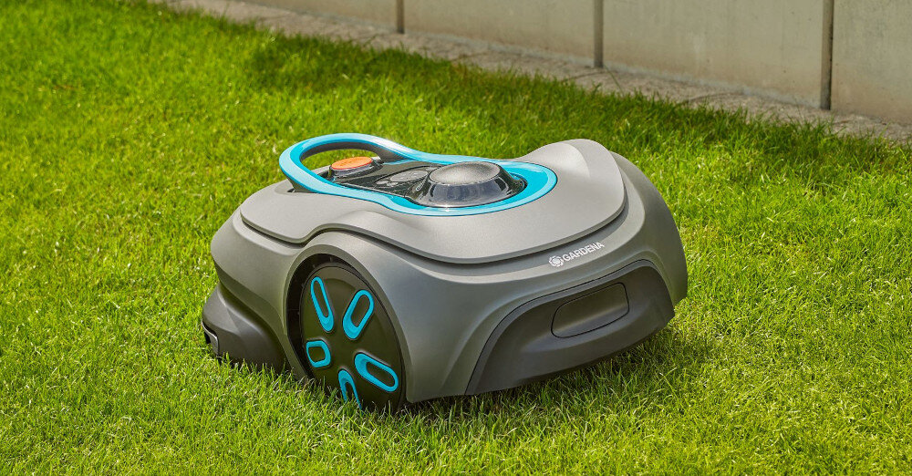 Na trawie znajduje się robot koszący w kolorze szarym z niebieskimi akcentami. Na obudowie widoczne jest logo \'GARDENA\'.