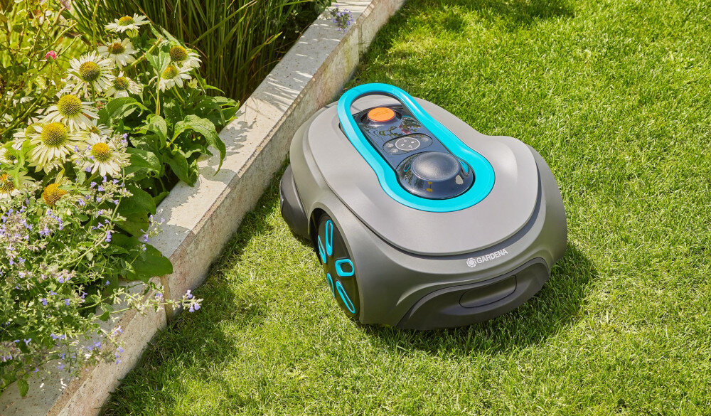 Na trawniku znajduje się robot koszący o szarej obudowie z niebieskimi akcentami. Na obudowie widoczne jest logo \'GARDENA\'.