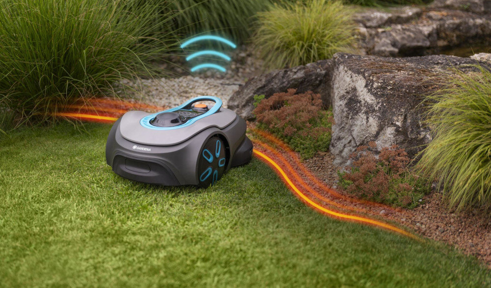 Robot koszący porusza się po trawniku obok kamieni i roślin, z widocznym logo Gardena. Nad robotem unosi się symbol sygnału Wi-Fi, a na trawie widoczna jest pomarańczowa linia prowadząca.