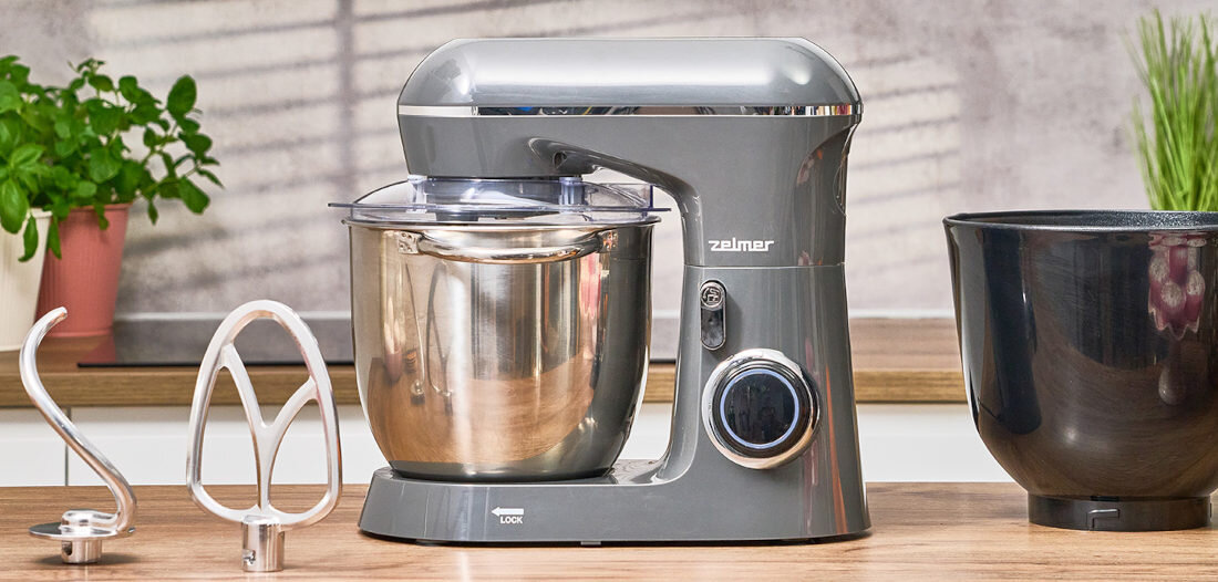 Na blacie kuchennym znajduje się szary robot kuchenny z metalową misą i logo \'zelmer\'. Obok robota leżą dwa mieszadła i dodatkowa czarna misa.