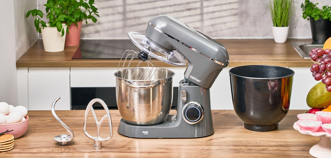 Na blacie kuchennym znajduje się robot kuchenny planetarny w kolorze szarym z metalową misą i trzema końcówkami do mieszania. Obok robota stoi dodatkowa czarna misa, a w tle widoczne są rośliny doniczkowe i owoce.