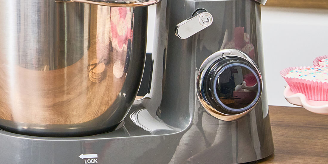 Na zdjęciu widoczny jest fragment robota kuchennego z metalową misą i pokrętłem. Obok znajduje się różowa foremka na babeczki z kolorową posypką.