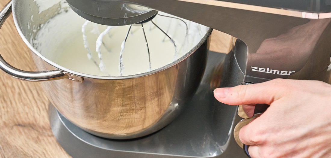 Robot kuchenny z metalową misą, w której znajduje się ubijana masa. Na urządzeniu widoczne jest logo \'zelmer\'.