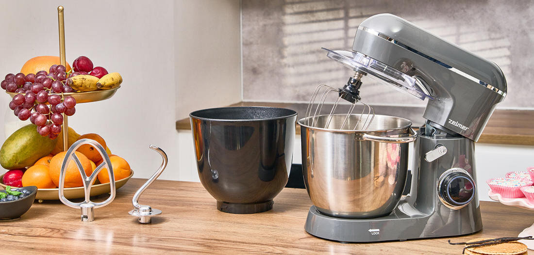 Na drewnianym blacie stoi robot kuchenny planetarny w kolorze szarym z logo \'zelmer\', obok którego znajdują się dwie misy – jedna metalowa, druga czarna. W tle widoczna jest patera z owocami oraz kilka akcesoriów kuchennych.