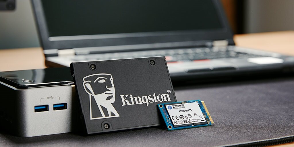 Na pierwszym planie znajduje się dysk SSD z logo Kingston, obok niego leży mniejszy moduł pamięci. W tle widoczny jest otwarty laptop.