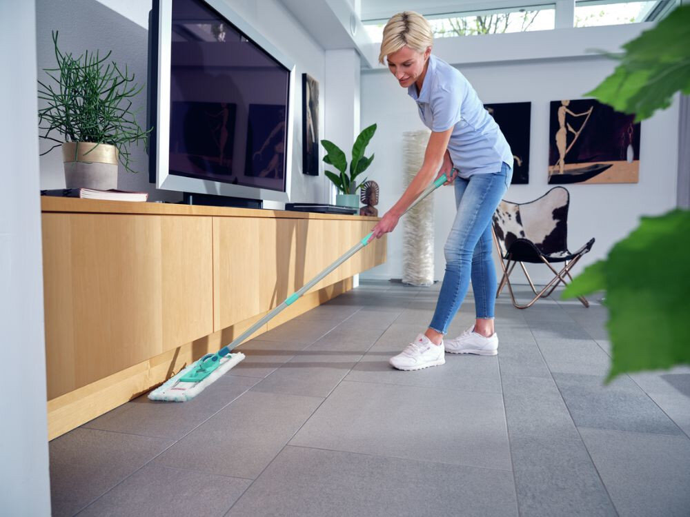 Osoba używa mopa do czyszczenia podłogi w nowoczesnym wnętrzu z drewnianą szafką i telewizorem. W tle widoczne są rośliny doniczkowe i obrazy na ścianie.