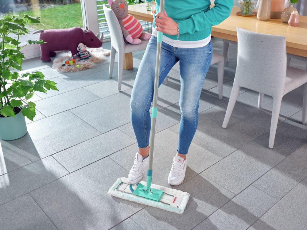 Osoba używa mopa z zielonym uchwytem na szarej podłodze w jasnym pomieszczeniu. W tle widać stół, krzesła i roślinę w doniczce.