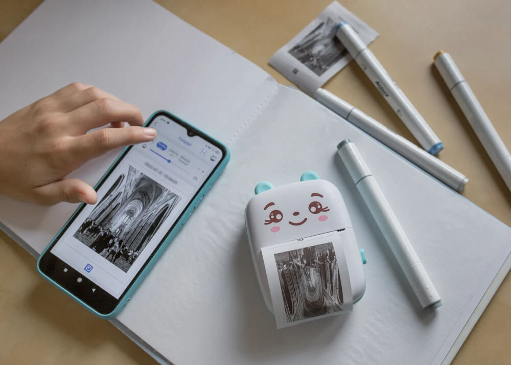 Na otwartej książce leży smartfon z wyświetlonym zdjęciem wnętrza katedry oraz mała drukarka z uśmiechniętą buźką, z której wychodzi wydruk tego samego zdjęcia. Obok znajdują się trzy długopisy i dodatkowy wydruk zdjęcia.