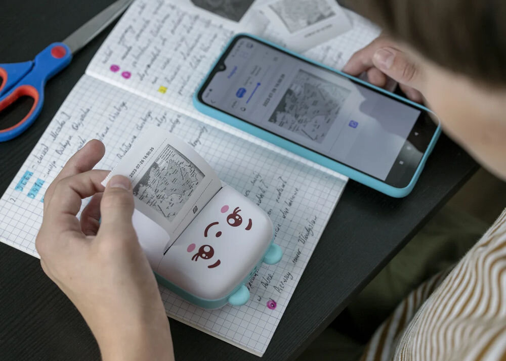 Osoba używa małej drukarki z uśmiechniętą buźką do drukowania obrazka na papierze termicznym. Obok znajduje się smartfon oraz zeszyt z notatkami na kratkowanym papierze.