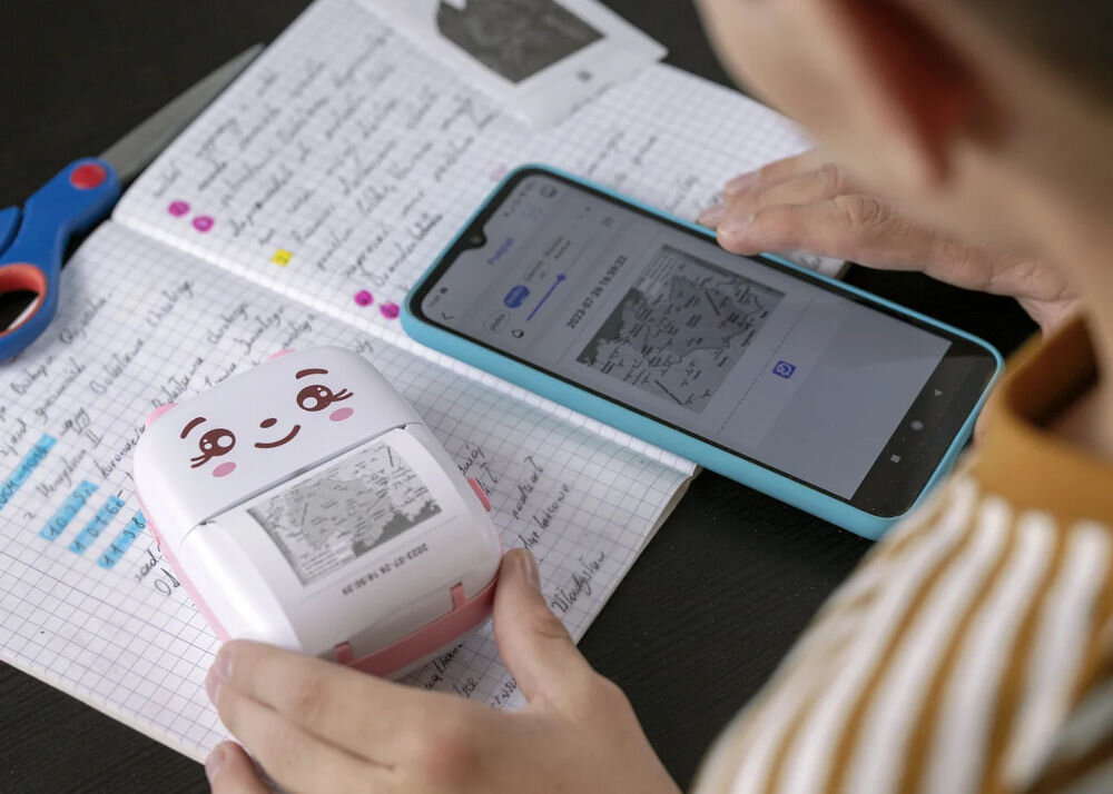 Na zdjęciu widać różową drukarkę z uśmiechniętą buźką, leżącą na zeszycie z notatkami. Obok znajduje się smartfon z wyświetlonym obrazem, a w tle widoczne są nożyczki.
