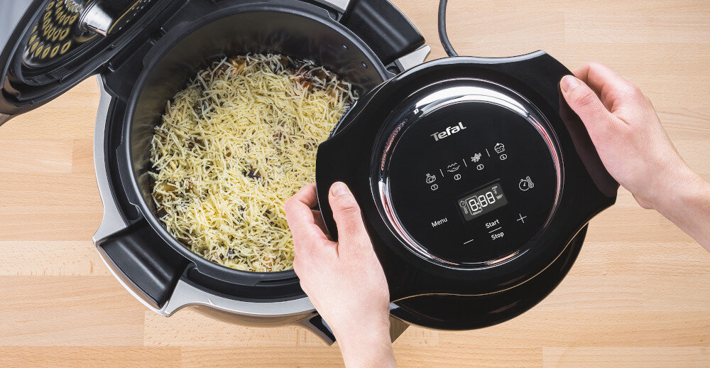 Osoba trzyma pokrywę z napisem \'Tefal\' nad urządzeniem kuchennym, w którym znajduje się potrawa posypana serem. Na pokrywie widoczne są ikony i wyświetlacz cyfrowy z napisem \'Menu\' oraz \'Start Stop\'.