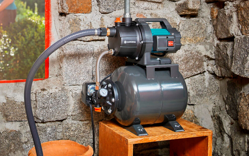 Na obrazie znajduje się hydrofor z widocznym logo \'GARDENA\', umieszczony na drewnianej podstawie przy kamiennej ścianie. Urządzenie ma ciemny zbiornik i węże podłączone do pompy.