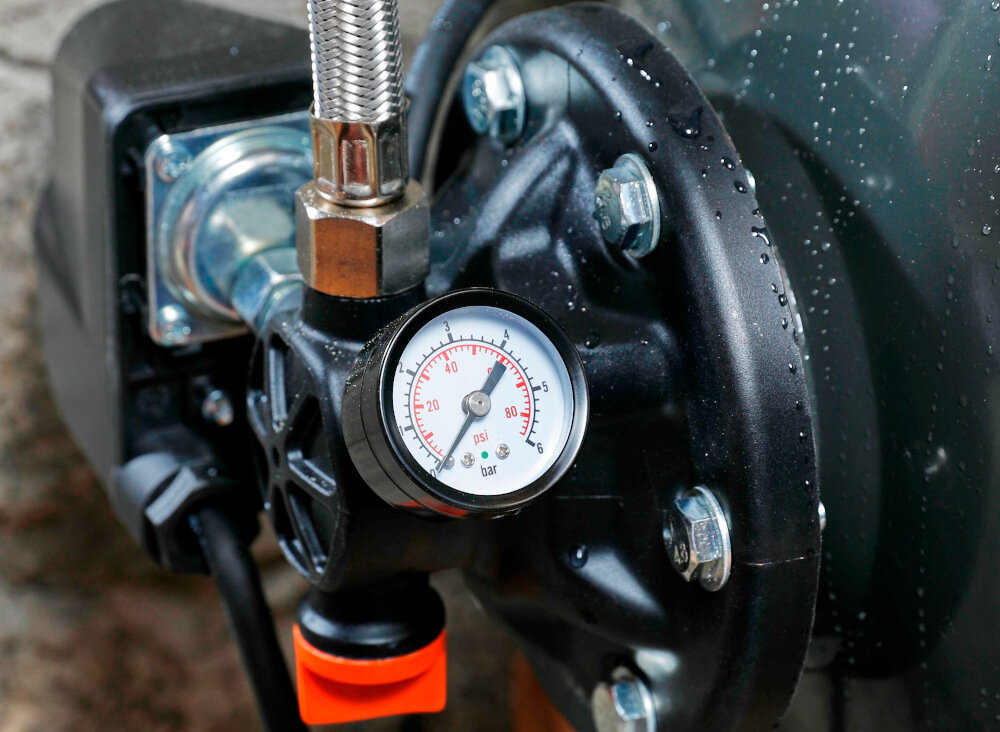 Zbliżenie na manometr z podziałką w barach i psi, zamontowany na czarnej obudowie urządzenia z metalowymi śrubami i przewodem. Na powierzchni widoczne są krople wody.