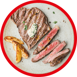 Na talerzu znajduje się kawałek grillowanego mięsa z masłem ziołowym oraz kilka kawałków pieczonych ziemniaków. Mięso jest częściowo pokrojone na plastry.