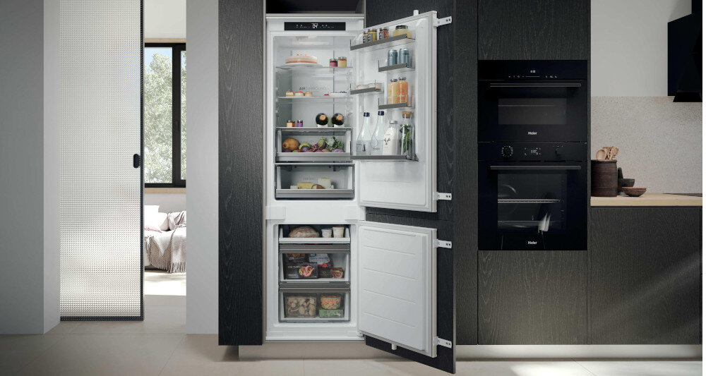 Otwarta lodówka w zabudowie kuchennej z widocznymi półkami wypełnionymi różnymi produktami spożywczymi. Obok znajduje się piekarnik i szafka kuchenna.