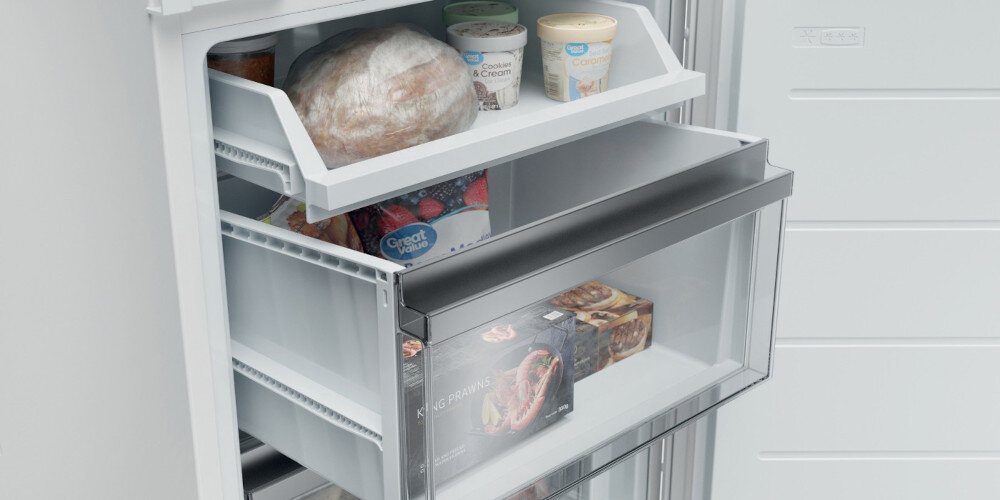 Wnętrze lodówki z widocznymi półkami i szufladami, wypełnione różnymi produktami spożywczymi, w tym opakowaniami lodów i mrożonek. Na jednej z półek znajduje się przezroczysta szuflada z napisem \'KING PRAWNS\'.