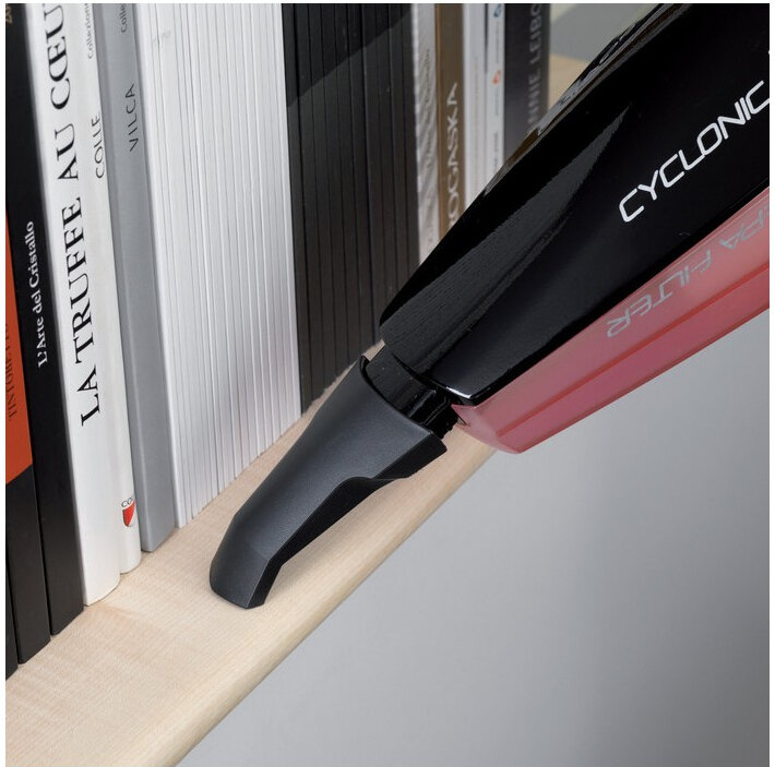 Czarny odkurzacz ręczny z napisem \'CYCLONIC\' i \'HEPA FILTER\' odkurza półkę z książkami. Widoczne są grzbiety książek z tytułami, m.in. \'LA TRUFFE AU CŒUR\'.