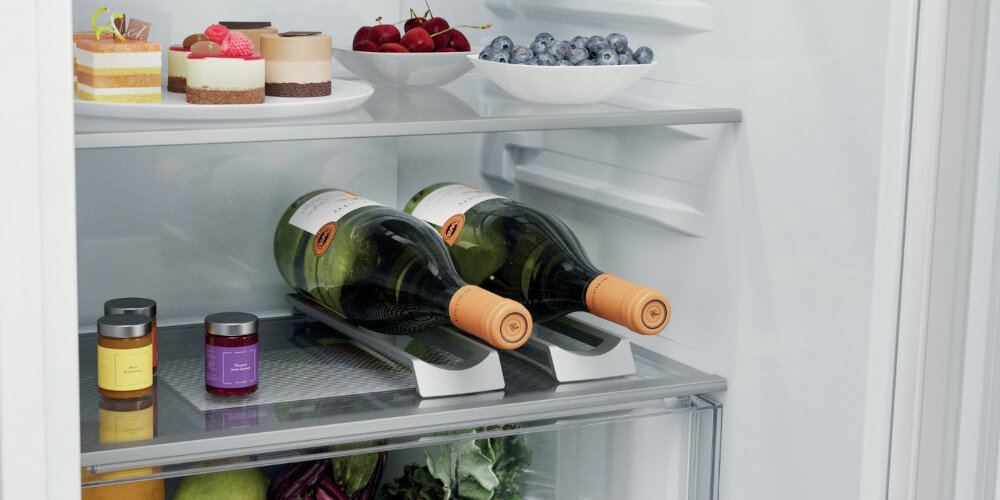 Wnętrze lodówki z widocznymi półkami, na których znajdują się ciasta, owoce, słoiczki oraz dwie butelki wina. Na dolnej półce widoczne są warzywa i owoce.