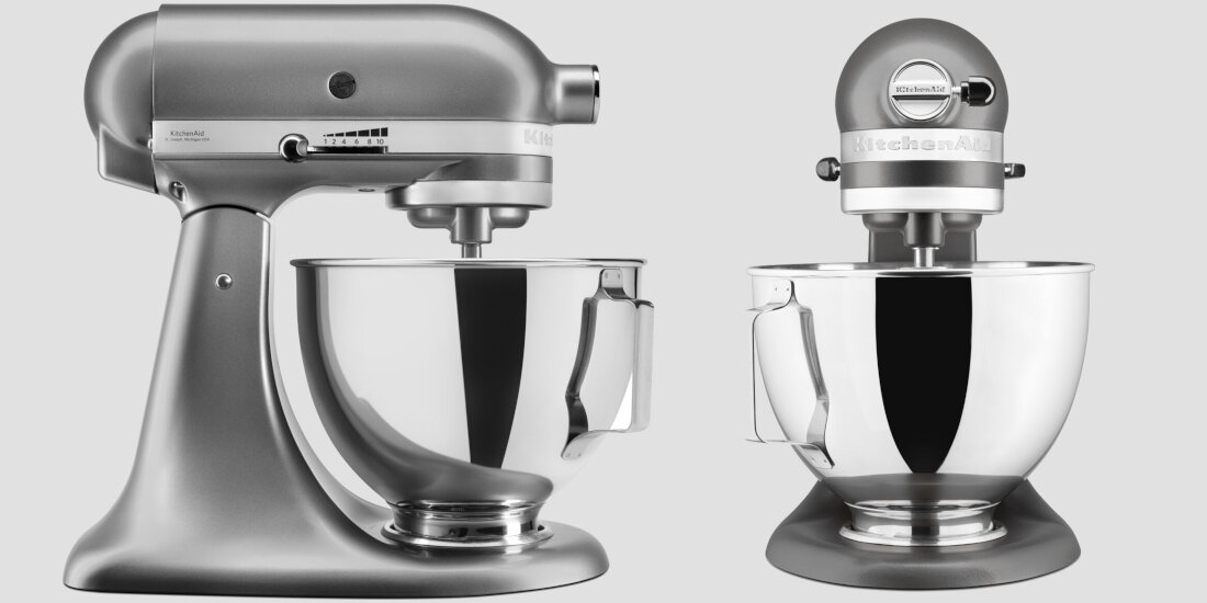 Na obrazie widoczne są dwa metaliczne roboty kuchenne z misami ze stali nierdzewnej. Na obu urządzeniach znajduje się logo \'KitchenAid\'.