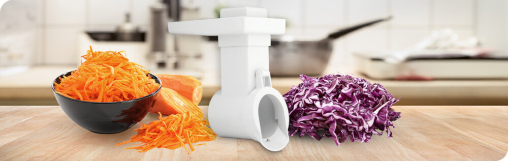 Na blacie kuchennym znajduje się biała maszynka do mięsa oraz miska z tartą marchewką i poszatkowana czerwona kapusta. W tle widoczna jest rozmyta kuchnia z garnkiem na kuchence.