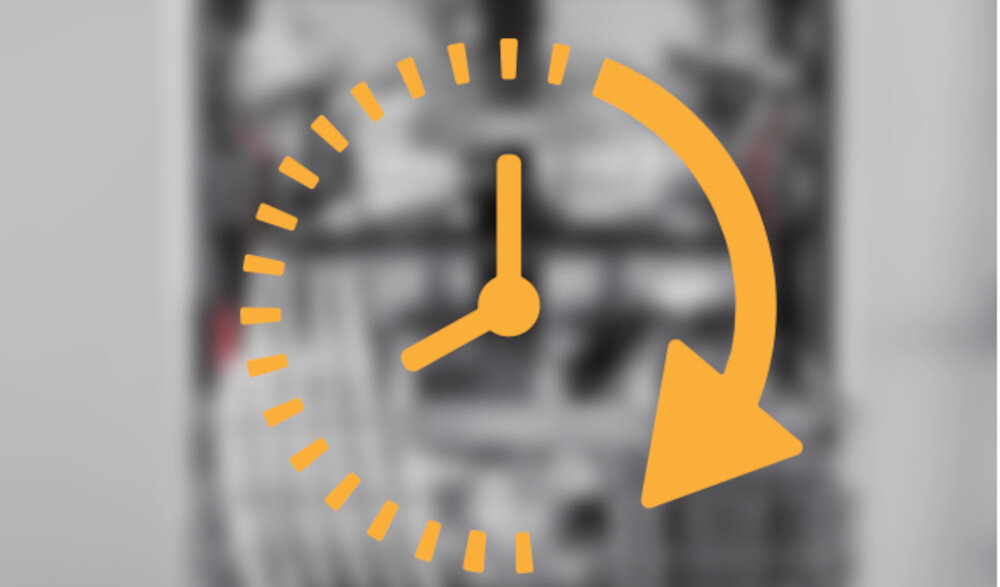 Obraz przedstawia ikonę zegara z pomarańczową strzałką skierowaną w dół i w prawo, na rozmytym tle wnętrza zmywarki.