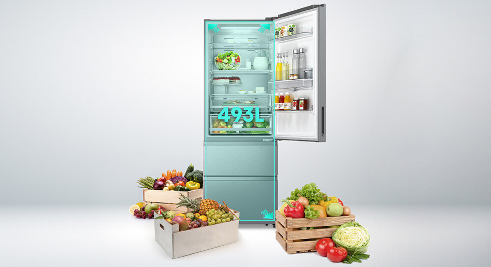 Otwarta lodówka z widocznymi półkami wypełnionymi jedzeniem i napojami, na drzwiach również znajdują się produkty. Obok lodówki stoją skrzynki z różnymi owocami i warzywami, a na lodówce widoczny jest napis \'493L\'.