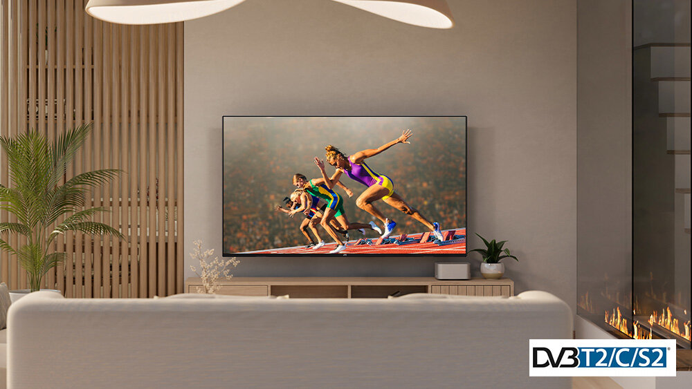 W salonie na ścianie wisi telewizor, na którym wyświetlany jest obraz sportowców na bieżni. W prawym dolnym rogu widoczny jest napis \'DVB T2/C/S2\'.