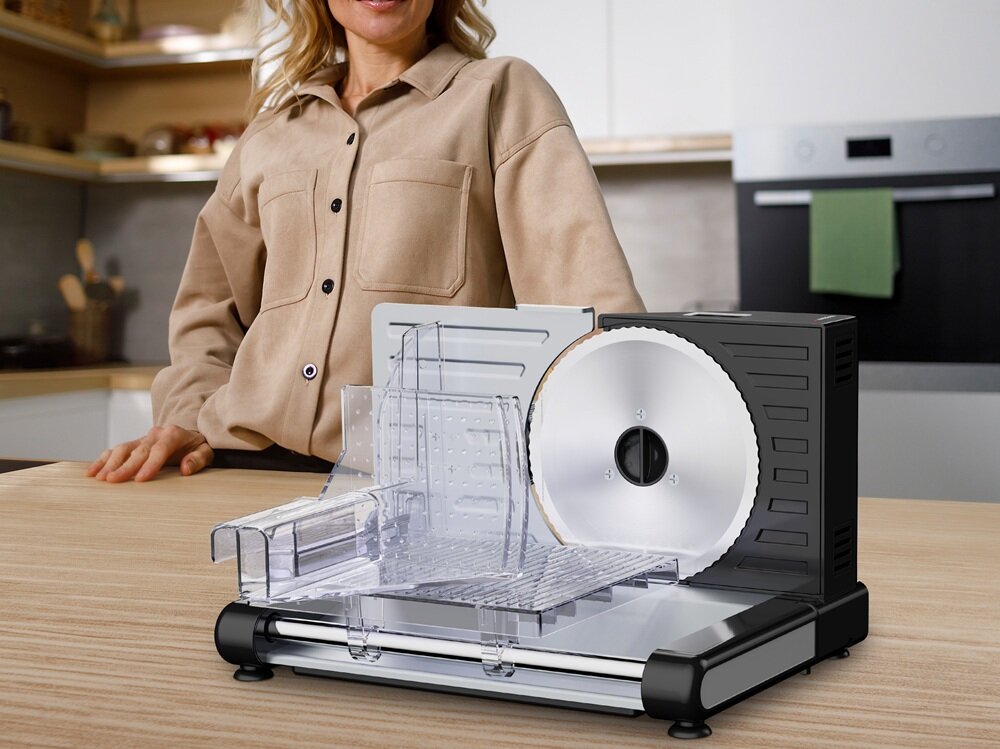 Na blacie kuchennym stoi krajalnica z przezroczystą osłoną i metalowym ostrzem. W tle widoczna jest osoba w beżowej koszuli.