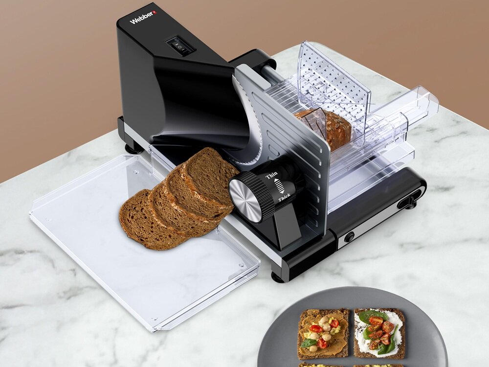 Na obrazie widoczna jest krajalnica z logo \'Webber\', na której krojony jest chleb. Obok znajduje się talerz z dwoma kromkami chleba z dodatkami.