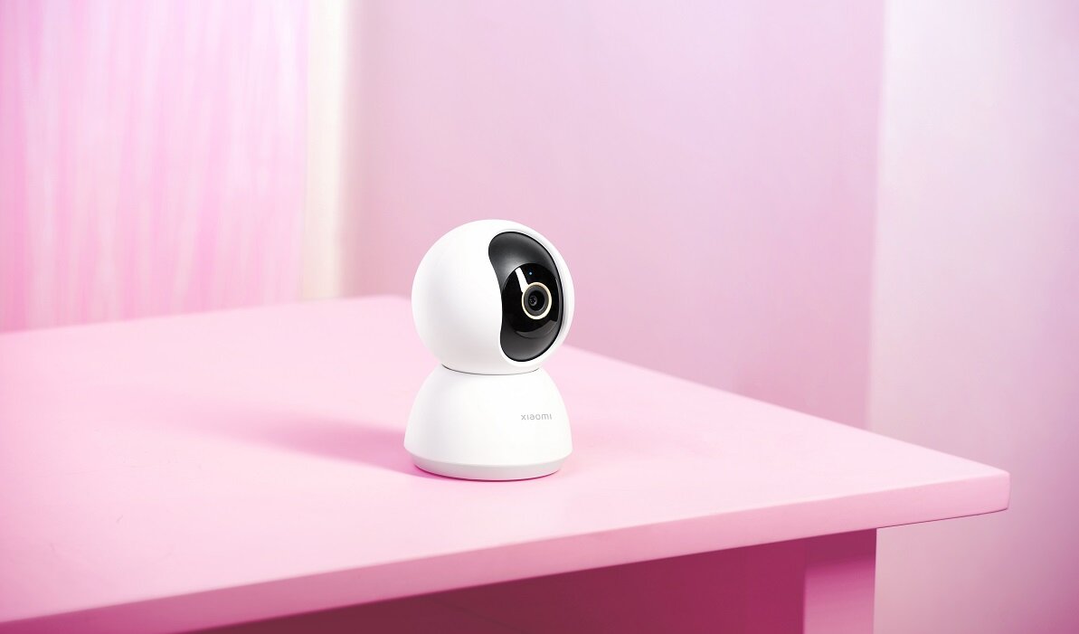Na różowym stole znajduje się biała, obrotowa kamera z czarnym obiektywem. Na obudowie widoczny jest napis \'XIAOMI\'.