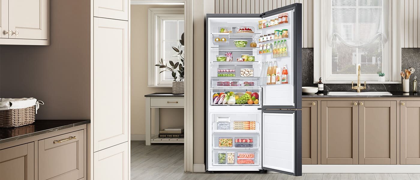 Otwarta lodówka w kuchni, wypełniona różnorodnymi produktami spożywczymi, takimi jak owoce, warzywa, napoje i nabiał. W tle widoczna jest elegancka kuchnia z jasnymi szafkami i dużym oknem.