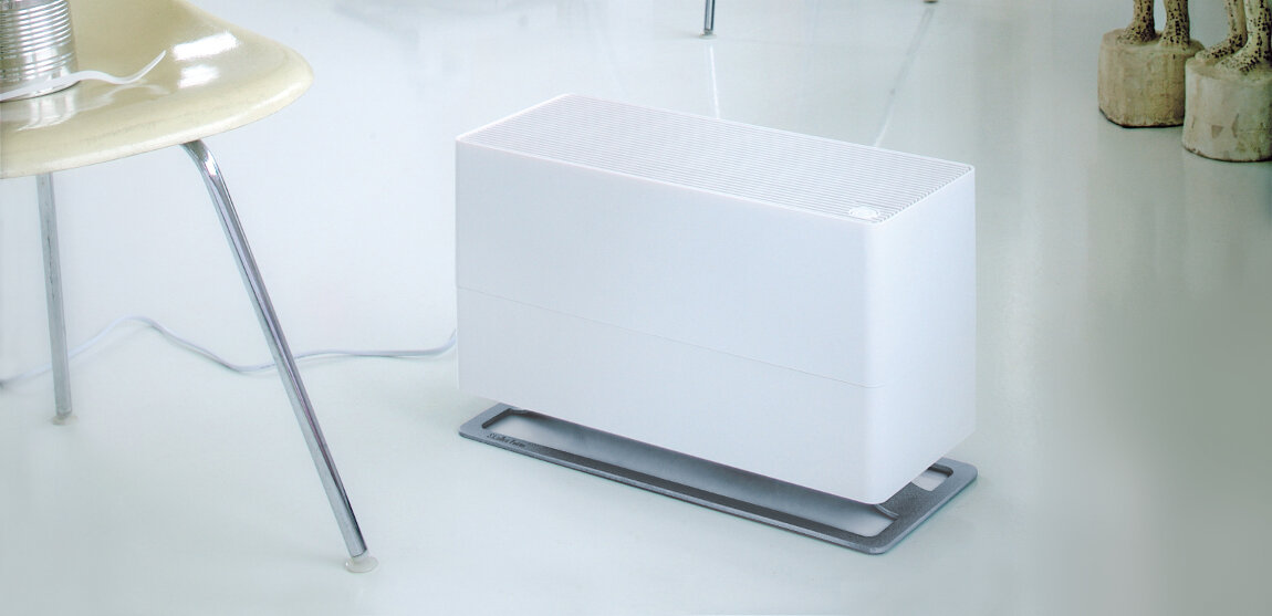 Na obrazie znajduje się biały nawilżacz powietrza o prostokątnym kształcie, umieszczony na podłodze. W tle widoczna jest część krzesła i elementy dekoracyjne.