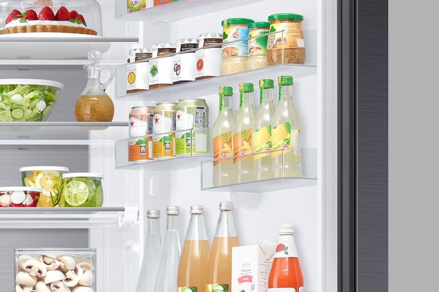 Wnętrze lodówki z półkami wypełnionymi różnymi produktami spożywczymi, takimi jak napoje w butelkach i puszkach, słoiki z przetworami oraz pojemniki z sałatkami. Na drzwiach lodówki znajdują się dodatkowe półki z napojami i słoikami.