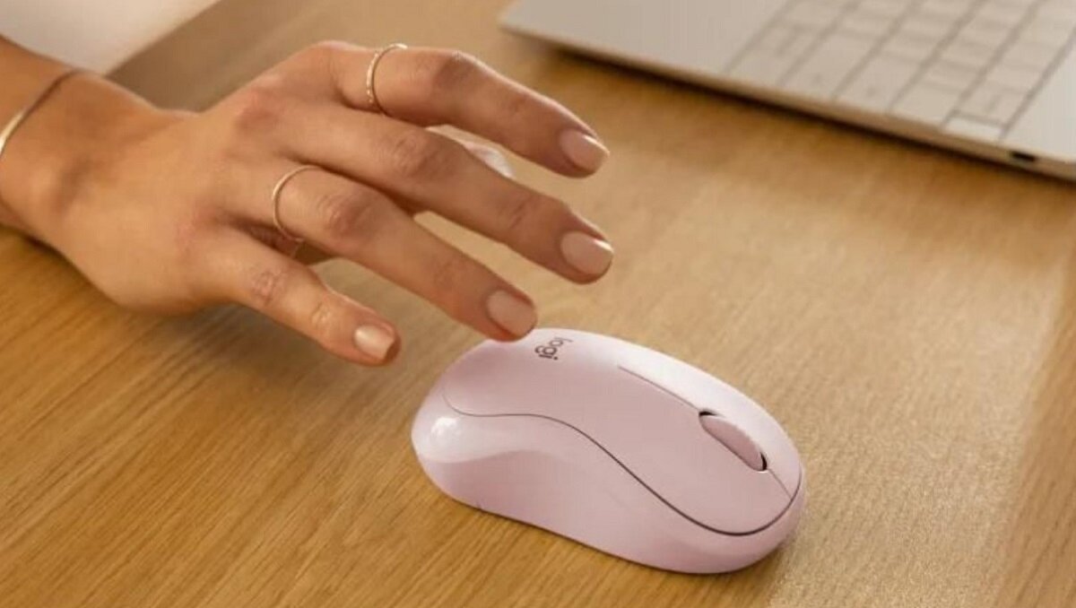 Na drewnianym blacie znajduje się biała mysz komputerowa z logo \'logi\'. Obok widoczna jest dłoń oraz fragment klawiatury.