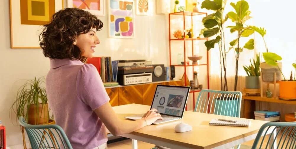 Osoba siedzi przy stole z laptopem, obok którego znajduje się biała mysz komputerowa. W tle widać rośliny doniczkowe i kolorowe dekoracje na ścianie.