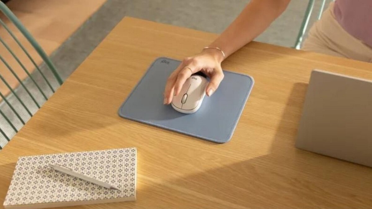 Na drewnianym stole znajduje się biała mysz komputerowa na niebieskiej podkładce, obok laptopa i notesu z długopisem. Mysz jest trzymana przez osobę, a na podkładce widoczne jest logo \'logi\'.