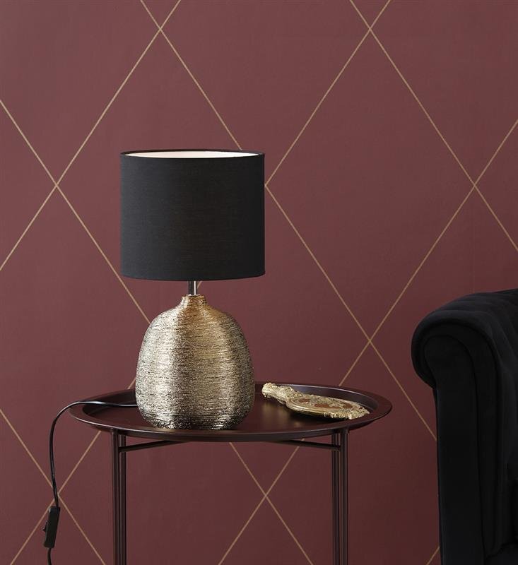 Na okrągłym stoliku znajduje się lampa stołowa z czarnym abażurem i złotą, teksturowaną podstawą. W tle widoczna jest bordowa ściana z geometrycznym wzorem.