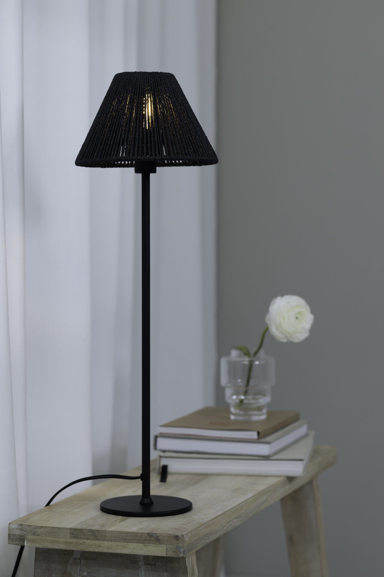 Czarna lampa stołowa z abażurem stoi na drewnianym stoliku, obok stosu książek i szklanej wazonu z białym kwiatem. W tle widoczna jest biała zasłona.