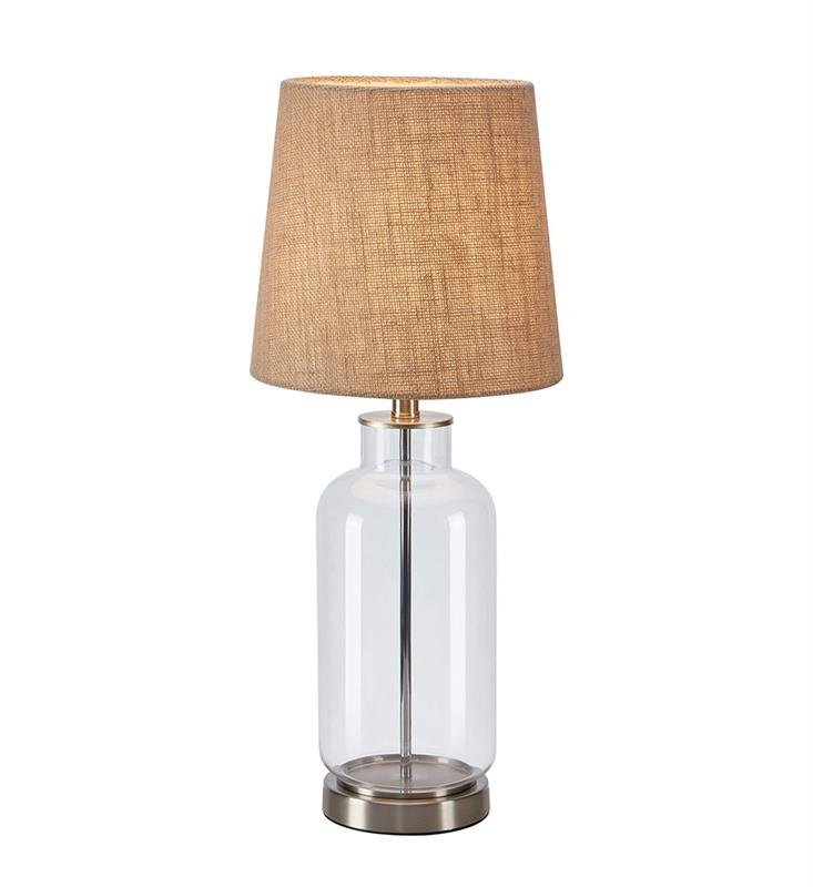 Lampa stołowa z przezroczystą szklaną podstawą i beżowym abażurem o teksturze przypominającej tkaninę. Podstawa lampy jest metalowa w kolorze srebrnym.